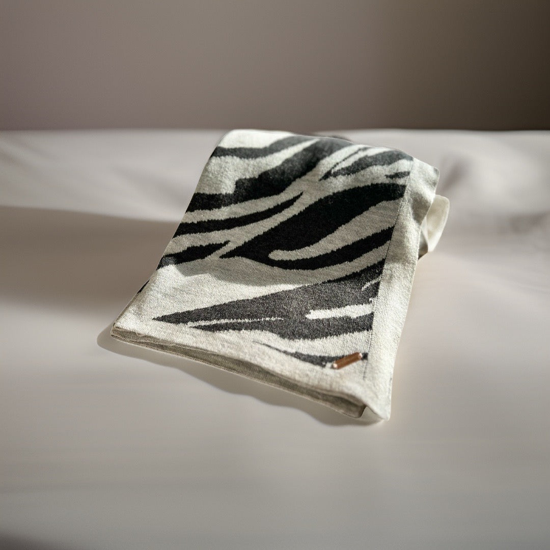 A folded luxury travel blanket with a zebra-like pattern in black and white, placed on a bed.